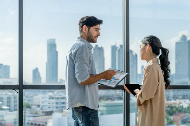 Un homme d'affaires professionnel présente des statistiques financières à un investisseur alors qu'il se tient près d'une fenêtre panoramique montrant un gratte-ciel ou une vue sur la ville Divers hommes d'affaires discutent du plan de marketing Tracery