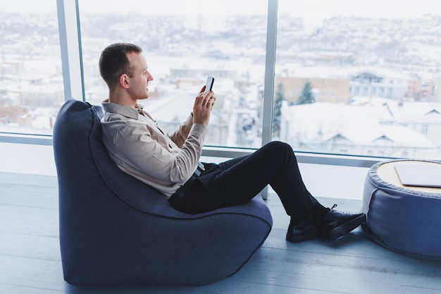 Homme d'affaires prospère assis dans un espace de travail moderne et vérifiant les notifications sur son téléphone portable, il porte une chemise et un pantalon