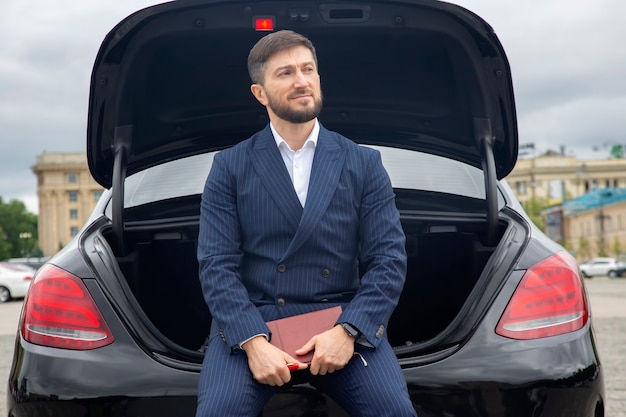 Un homme d'affaires prospère est assis avec un journal sur le capot de sa voiture prestigieuse