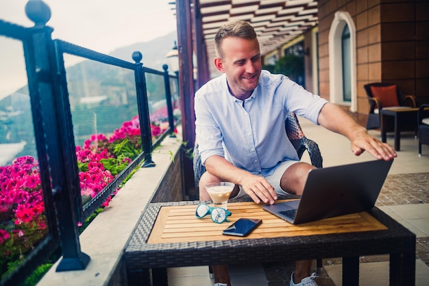 Un homme d'affaires prospère est assis à une table avec un ordinateur portable, il travaille en vacances Vacances et travail à distance Le gars est assis dans un restaurant sur une terrasse avec vue panoramique