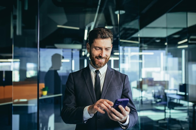 Homme d'affaires prospère et heureux avec une barbe utilise un téléphone portable sourit et se réjouit regarde l'écran du smartphone dans un bureau moderne