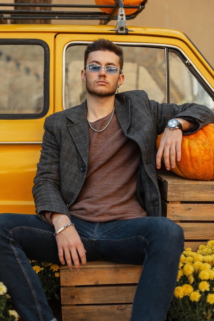 Homme d'affaires prospère à la mode avec une coiffure et des lunettes de soleil dans des vêtements décontractés avec un jean manteau et une montre est assis près d'une vieille voiture rétro jaune avec une citrouille et des fleurs en plein air