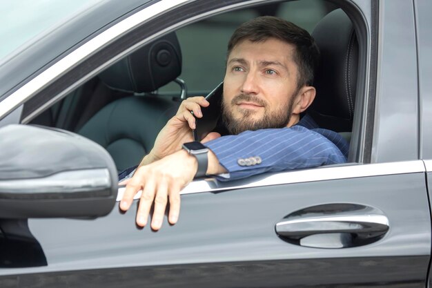 Un homme d'affaires prospère avec un téléphone à la main est assis au volant d'une voiture prestigieuse