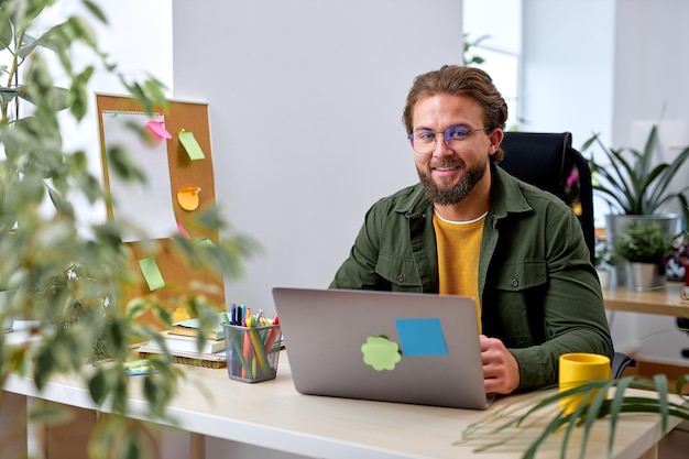 Homme d'affaires prospère travaillant sur ordinateur portable au bureau. Homme d'affaires assis au bureau travaillant sur un nouveau projet, souriant, se sentant heureux. beau mec barbu caucasien en tenue décontractée. notion de réussite