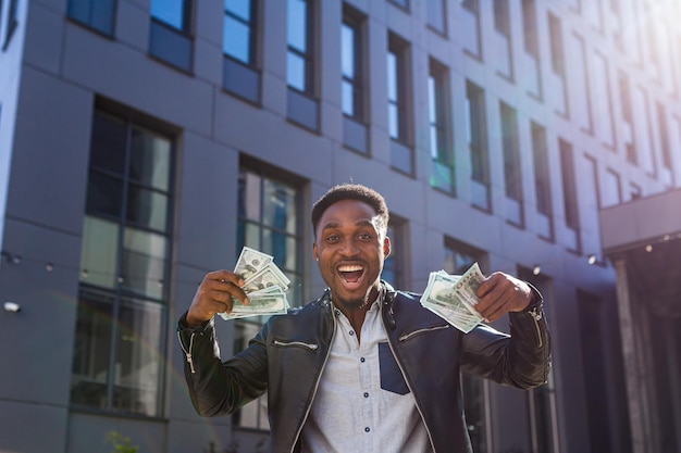 Un homme d'affaires prospère en vêtements décontractés se réjouit du succès, l'Afro-américain regarde dans la caméra, sourit et détient des dollars en espèces