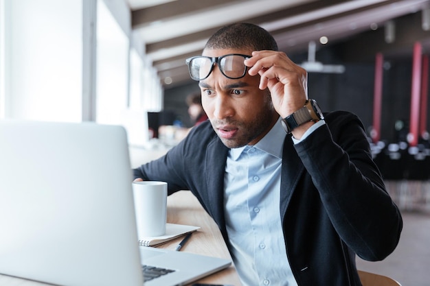 Photo homme d'affaires recevant des nouvelles négatives, touchant ses lunettes, étant fou, contrarié et surpris devant un ordinateur portable