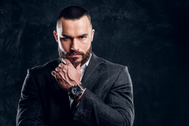 Un homme d'affaires sceptique sérieux avec une barbe et des vêtements sombres se tient dans un studio photo sombre.