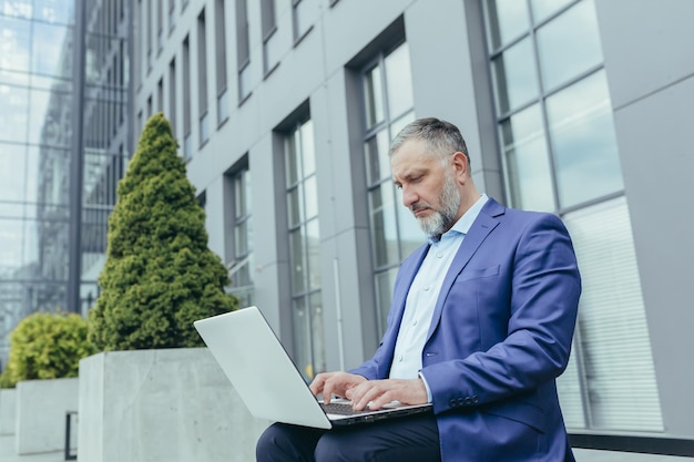 Homme d'affaires senior aux cheveux gris sérieux et concentré à l'extérieur de l'immeuble de bureaux travaillant à l'aide d'un ordinateur portable mâle