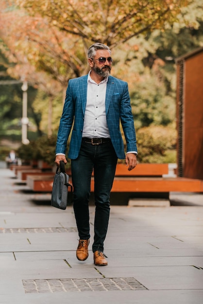 Un homme d'affaires senior en costume et lunettes de soleil marchant dans la ville tout en portant un sac.