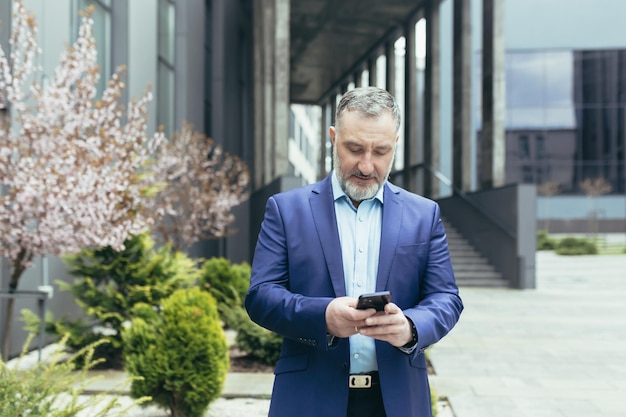 Homme d'affaires senior expérimenté aux cheveux gris à l'extérieur du bureau souriant et heureux en utilisant la lecture du patron du téléphone