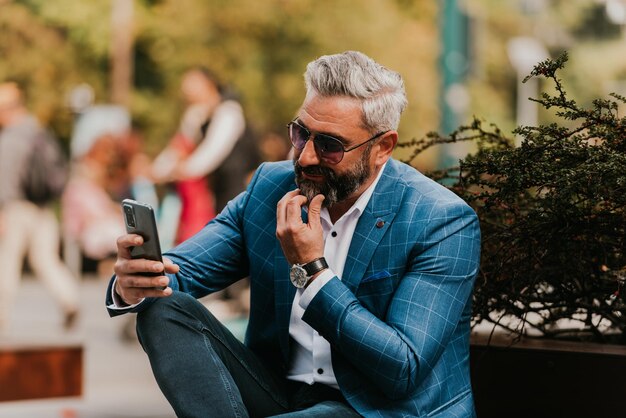 Homme d'affaires senior moderne utilisant un smartphone pour une réunion en ligne assis sur un banc.