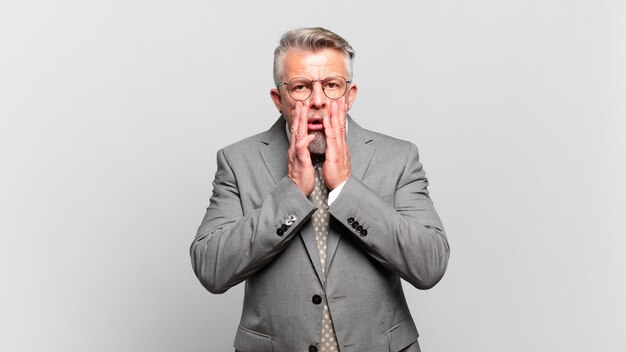 Homme d'affaires senior se sentant choqué et effrayé, l'air terrifié avec la bouche ouverte et les mains sur les joues