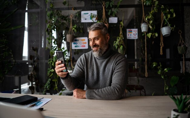 Homme d'affaires senior travaillant au téléphone portable et souriant à la caméra au bureau contemporain senior