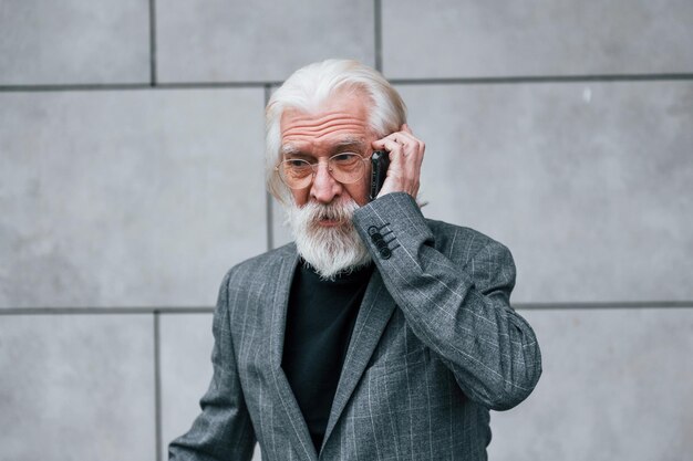 Un homme d'affaires senior en vêtements formels aux cheveux gris et à la barbe parle à l'extérieur par téléphone