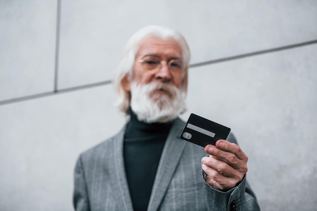 Un homme d'affaires senior en vêtements formels avec des cheveux gris et une barbe est à l'extérieur détient une carte de crédit