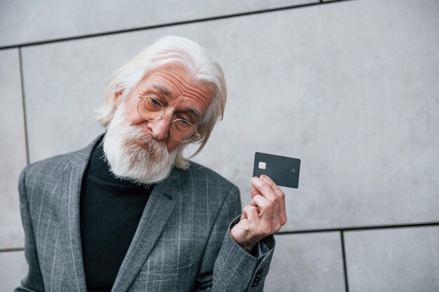 Un homme d'affaires senior en vêtements formels avec des cheveux gris et une barbe est à l'extérieur détient une carte de crédit