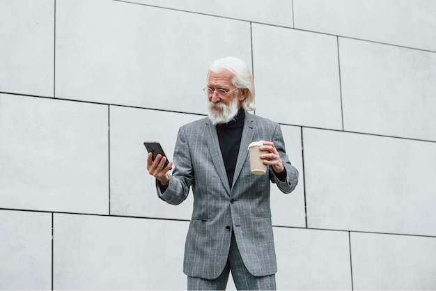 Un homme d'affaires senior en vêtements formels avec des cheveux gris et une barbe est à l'extérieur avec un téléphone et une tasse de boisson