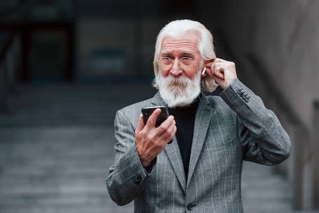 Homme d'affaires senior en vêtements formels avec des cheveux gris et une barbe est à l'extérieur en train d'essayer des écouteurs sans fil avec téléphone