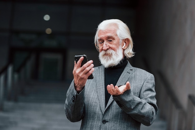 Homme d'affaires senior en vêtements formels avec des cheveux gris et une barbe est à l'extérieur en train d'essayer des écouteurs sans fil avec téléphone