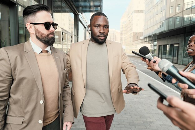 Homme d'affaires sérieux en lunettes de soleil avec son garde du corps donnant une interview aux journalistes de la ville