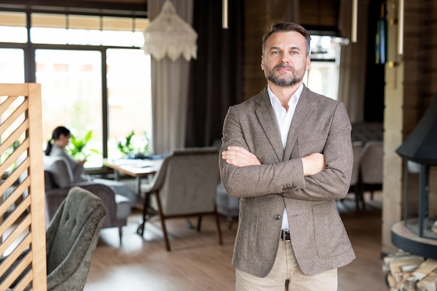 Homme d'affaires sérieux prospère en tenue de soirée en croisant les bras sur la poitrine en se tenant debout devant la caméra au café