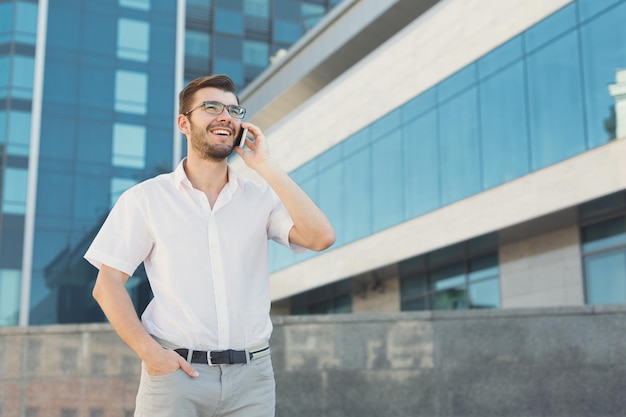 Un homme d'affaires souriant et attrayant dans des lunettes parle sur un téléphone portable et met sa main dans une poche devant un immeuble de bureaux moderne, copiez l'espace. Concept d'entreprise et de communication