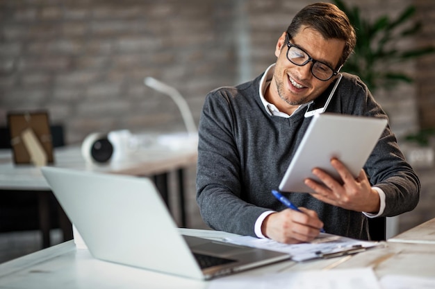 Homme d'affaires souriant ayant une journée bien remplie au travail Il parle au téléphone tout en utilisant le pavé tactile et en prenant des notes sur le papier