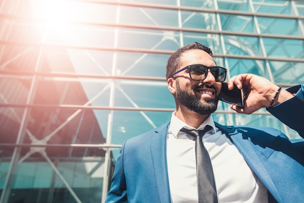 Un homme d'affaires souriant barbu porte un costume bleu et des lunettes de soleil parlant via un smartphone devant un bâtiment moderne