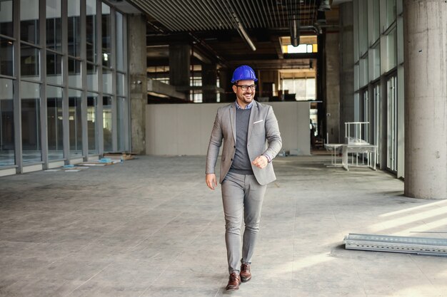 Homme d'affaires souriant avec casque sur la tête marchant à travers le chantier de construction de son immeuble.