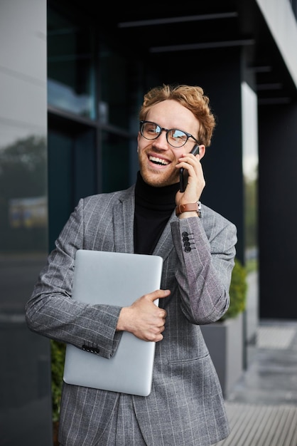 Homme d'affaires souriant portant un costume élégant, lunettes parlant sur un téléphone portable marchant dans la rue