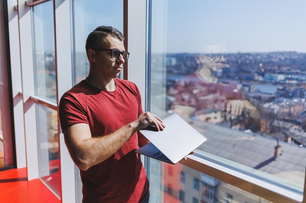 Un homme d'affaires souriant se tient près de la fenêtre tout en travaillant sur un ordinateur portable Concept multitâche Un pigiste occupé à lunettes tient un ordinateur portable dans ses mains tout en se tenant près de la fenêtre