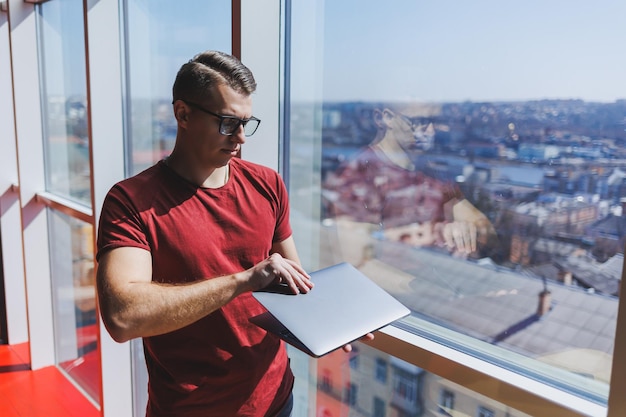 Un homme d'affaires souriant se tient près de la fenêtre tout en travaillant sur un ordinateur portable Concept multitâche Un pigiste occupé à lunettes tient un ordinateur portable dans ses mains tout en se tenant près de la fenêtre