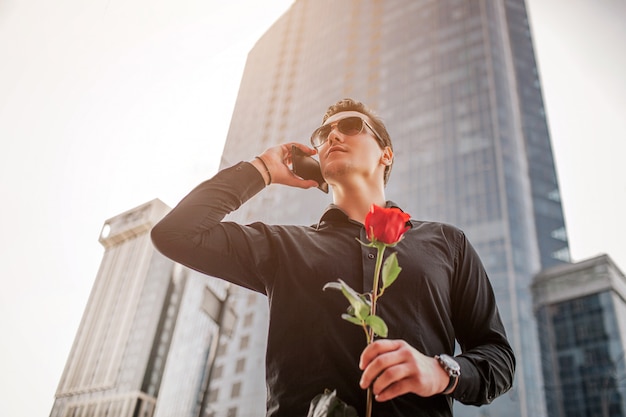 Homme affaires, stand, devant, gratte-ciel, conversation, téléphone Il tenait une rose rouge à la main. Le soleil brille dehors.