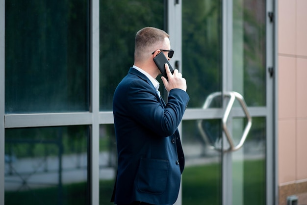 Un homme d'affaires avec un téléphone dans les mains se tient devant le bâtiment de l'entreprise et parle au téléphone