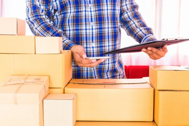 Homme d'affaires travaillant avec un téléphone portable et l'emballage de la boîte de colis brune à la maison. le vendeur de mains prépare le produit prêt à livrer au client. Vente en ligne, commerce électronique Démarrage du concept d'expédition.