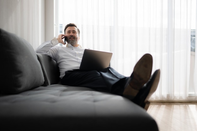 Homme d'affaires utilisant un ordinateur portable et parlant au téléphone alors qu'il est assis sur le canapé à la maison