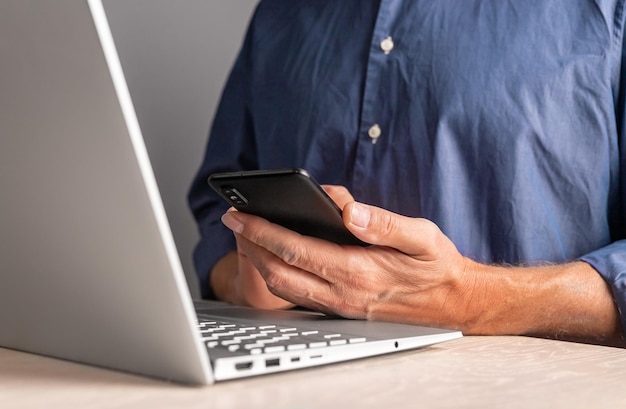 Homme d'affaires utilisant un smartphone pour le travail ou les loisirs au bureau à la maison, assis à table avec un ordinateur portable et tenant un téléphone