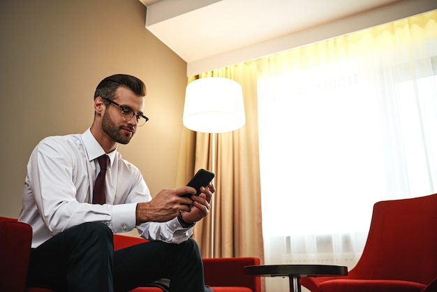 Homme d'affaires avec valise et smartphone assis sur un canapé dans le hall de l'hôtel