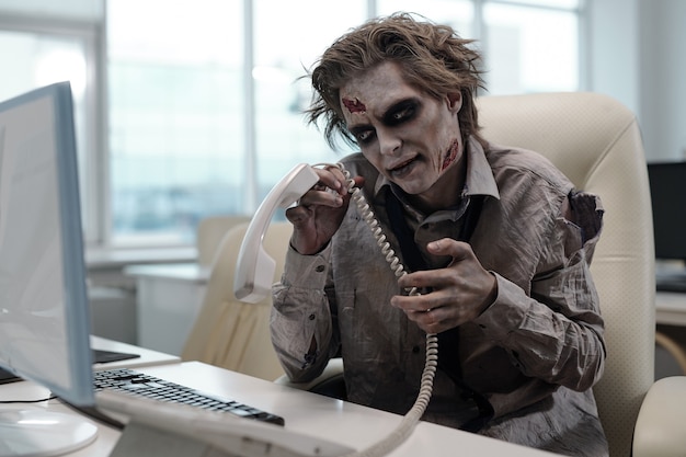 Homme d'affaires de zombie avec récepteur de téléphone assis devant l'ordinateur