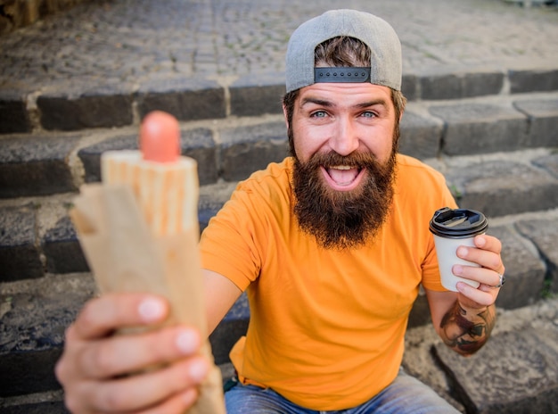 Homme affamé collation Guy mangeant un hot-dog Homme barbu morsure savoureuse saucisse et boisson gobelet en papier Nourriture de rue si bonne Mode de vie urbain nutrition Malbouffe Hipster insouciant mange de la malbouffe assis dans les escaliers