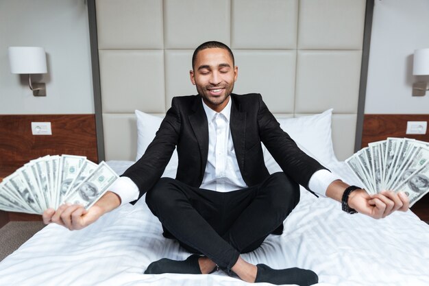 Homme africain en costume médite avec de l'argent dans les mains et les yeux fermés sur le lit dans la chambre d'hôtel