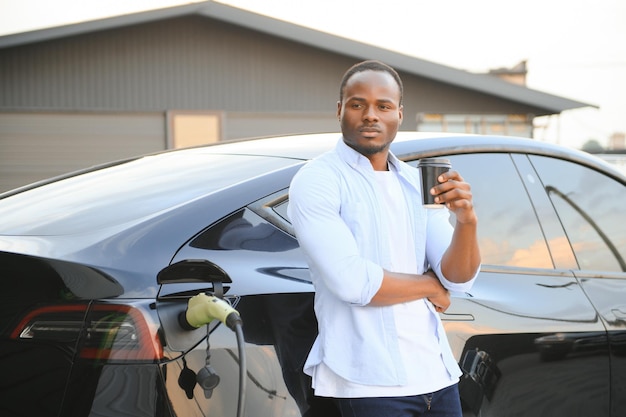 Homme africain debout près d'une voiture électrique avec un câble de charge dans la prise Charge de véhicule écologique