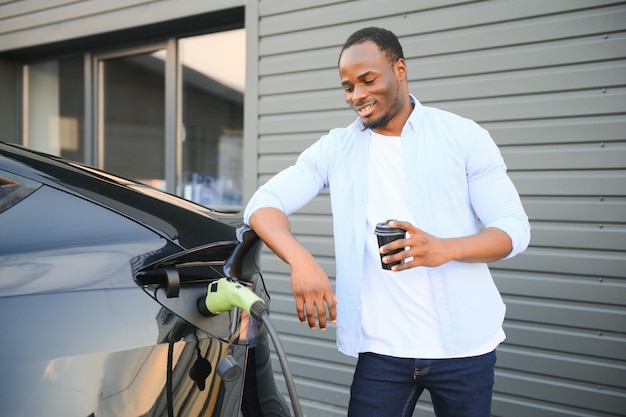 Un homme africain élégant avec une tasse de café à la main insère une prise dans la prise de chargement de la voiture électrique