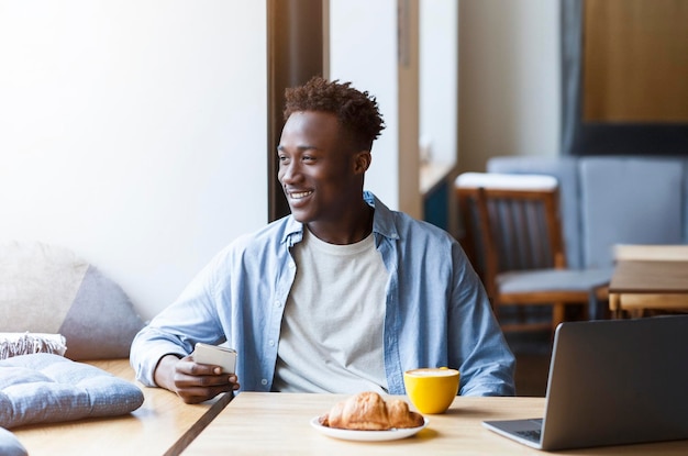 Homme africain millénaire avec smartphone et ordinateur portable assis dans un café