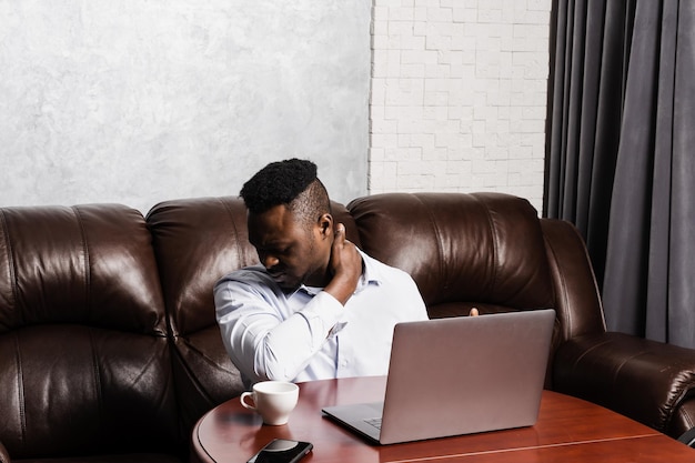 Un homme africain avec un ordinateur portable ressent des douleurs au cou et à la colonne vertébrale à cause de la compression des nerfs rachidiens L'ostéochondrose de la colonne cervicale est un syndrome radiculaire après un long travail à l'ordinateur