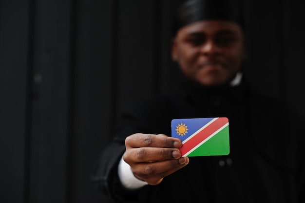 Homme africain porter durag noir tenir le drapeau de la Namibie à la main mur sombre isolé.