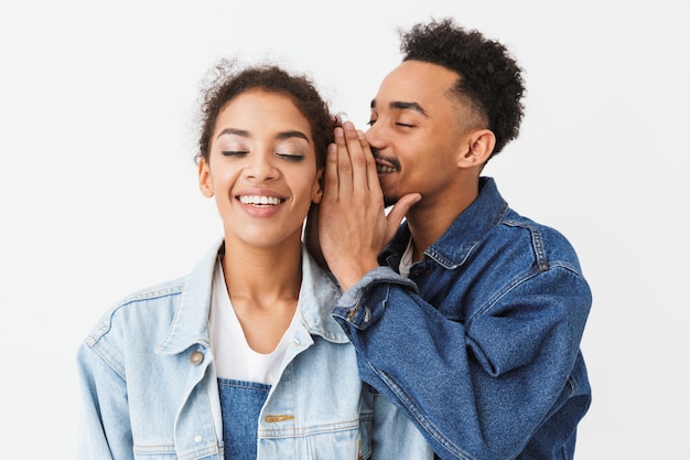 Homme africain souriant, parler quelque chose à l'oreille de sa petite amie heureuse qui écoute les yeux fermés sur mur gris