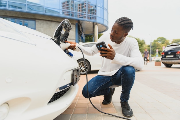 Homme africain tenant un câble de charge à portée de main, debout près d'une voiture électrique de luxe.