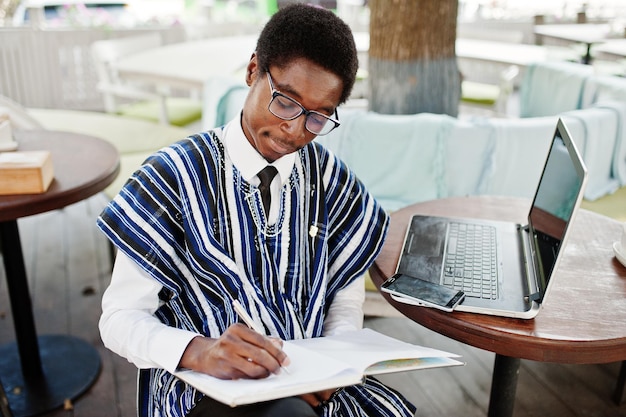 Homme africain en vêtements traditionnels et lunettes assis derrière un ordinateur portable au café en plein air et écrivant quelque chose sur son cahier
