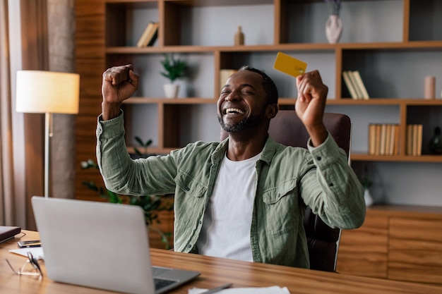 Un homme afro-américain adulte excité et satisfait se réjouit de gagner du cashback et des achats en ligne